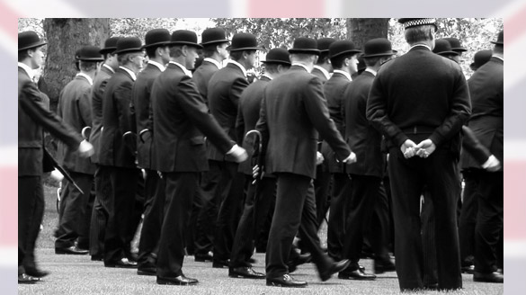 English veterans march in formation, wearing suits and derbies while clutching umbrellas as a Bobby looks on